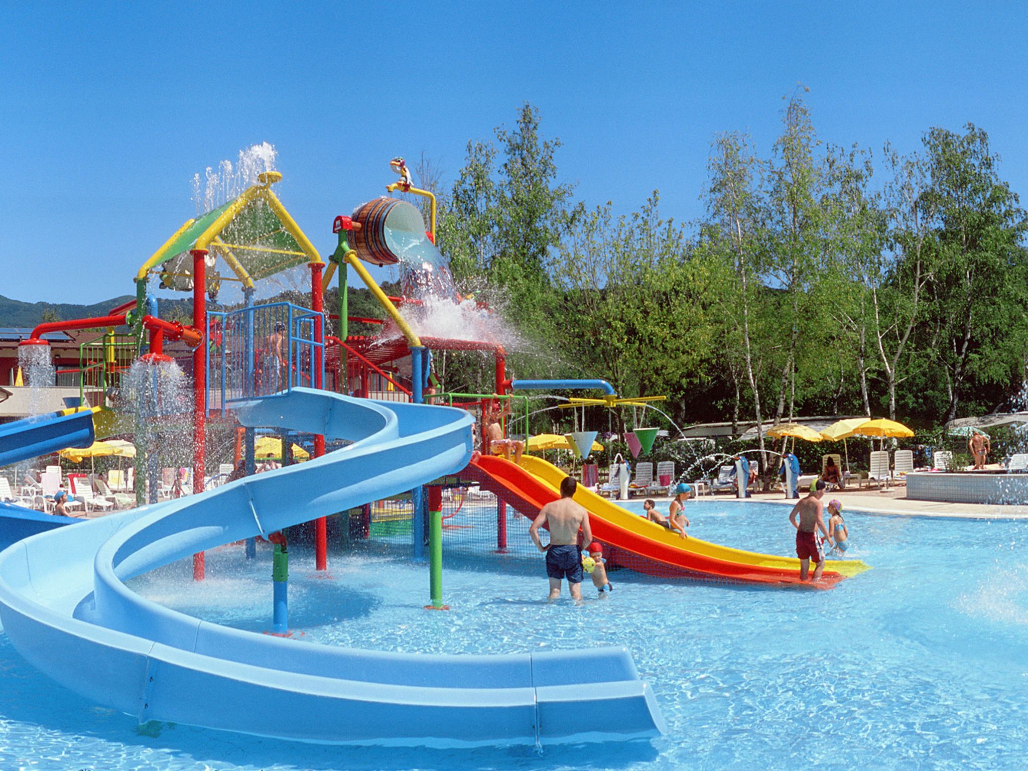 Parque acuático con toboganes y atracciones acuáticas, rodeado de árboles y una zona de piscina con sillas de descanso.