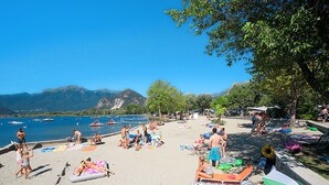 A beach with people sunbathing and swimming, boats in the water, and mountains in the background. - Fon 105 by Interhome (Fondotoce)