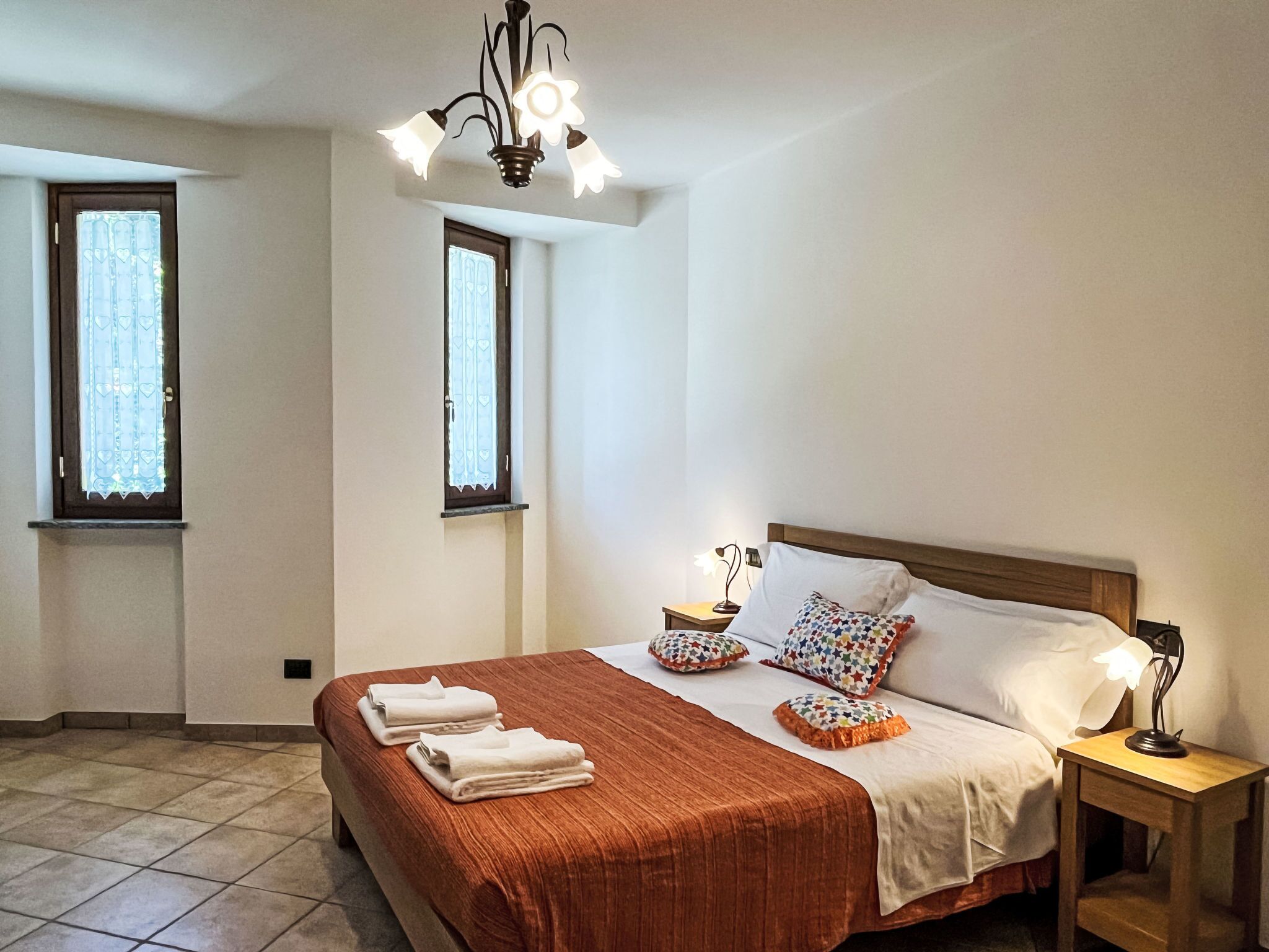 Ein Schlafzimmer mit einem großen Bett, zwei Nachttischen, einer Chandelüster und zwei Fenstern.