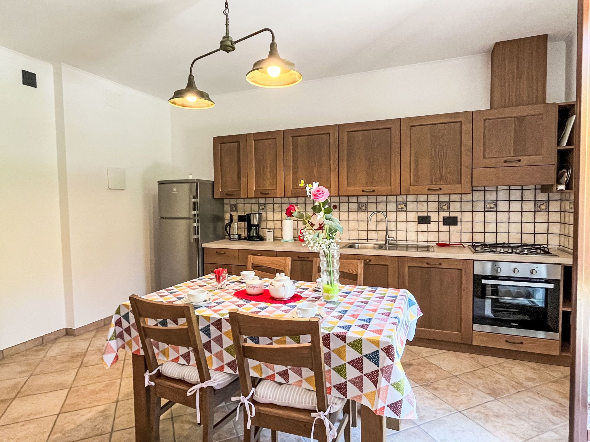 Une cuisine avec des meubles en bois, un plan de travail recouvert de carreaux, un réfrigérateur en acier inoxydable, une cuisinière à gaz et une table à manger avec des chaises.