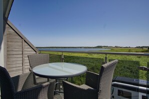 Eine Terrasse mit Korbmöbeln und einem Glastisch mit Blick auf ein Gewässer.
