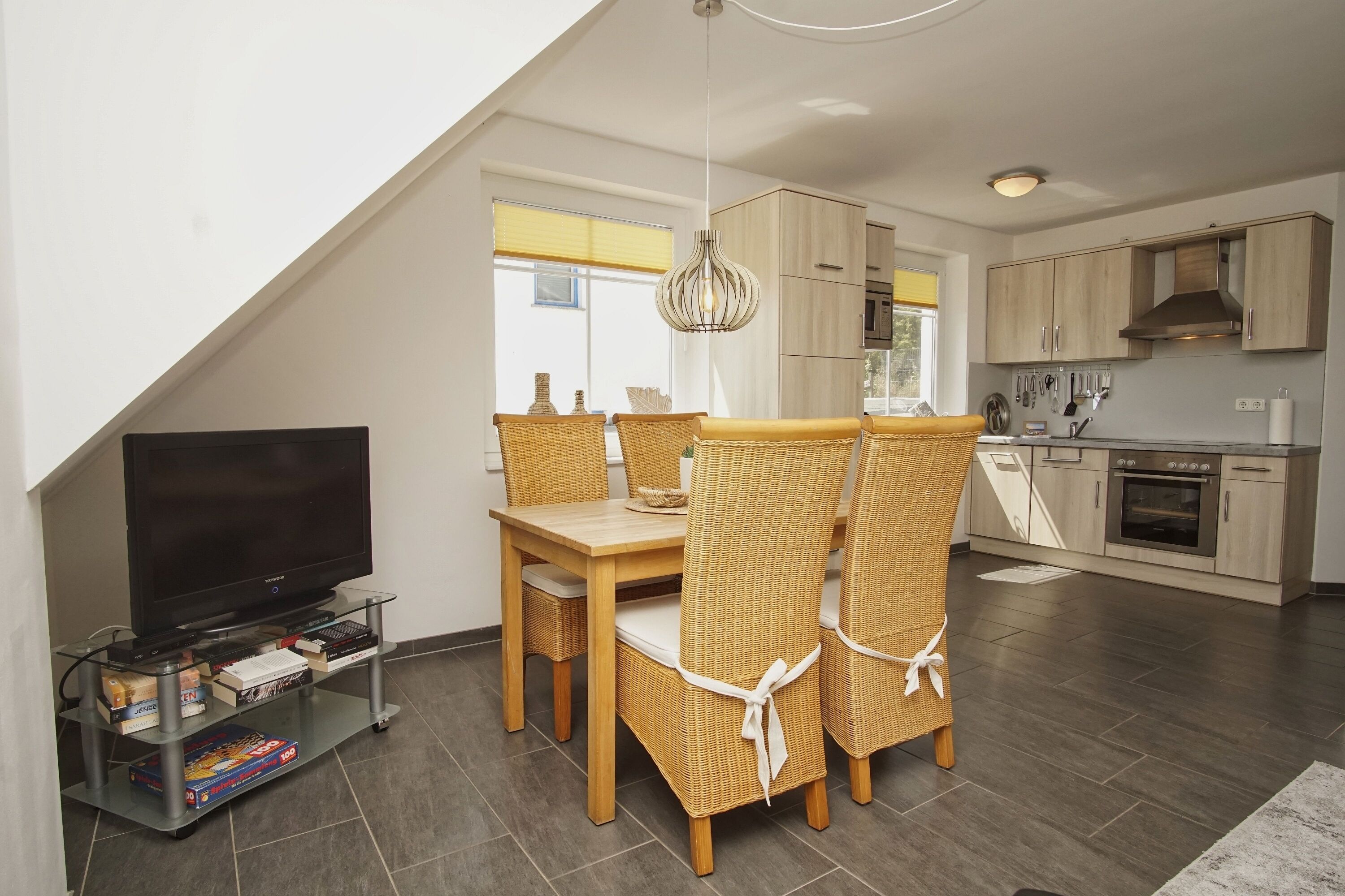 Een moderne keuken met houten kastjes, een inbouwoven en een eethoek met rieten stoelen en een glazen tafel.