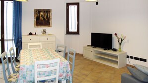 Un coin repas avec une table recouverte d’une nappe à motifs floraux, entourée de chaises. Un téléviseur est posé sur un meuble à droite, et un vase rempli de fleurs se trouve également sur ce meuble.