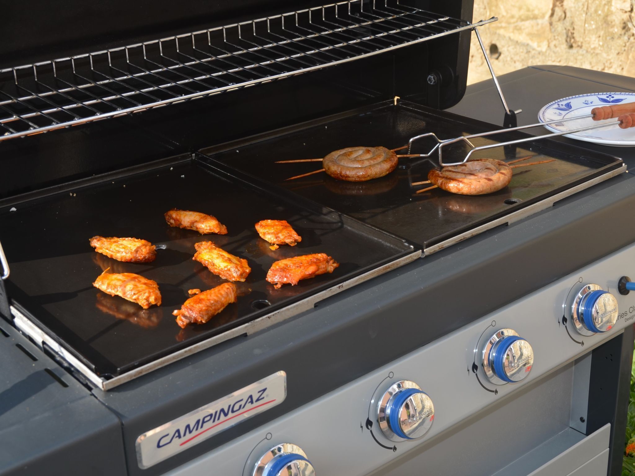 Un réchaud de camping portable, équipé d’une plaque de cuisson, sur lequel on fait griller des ailes de poulet et des saucisses.