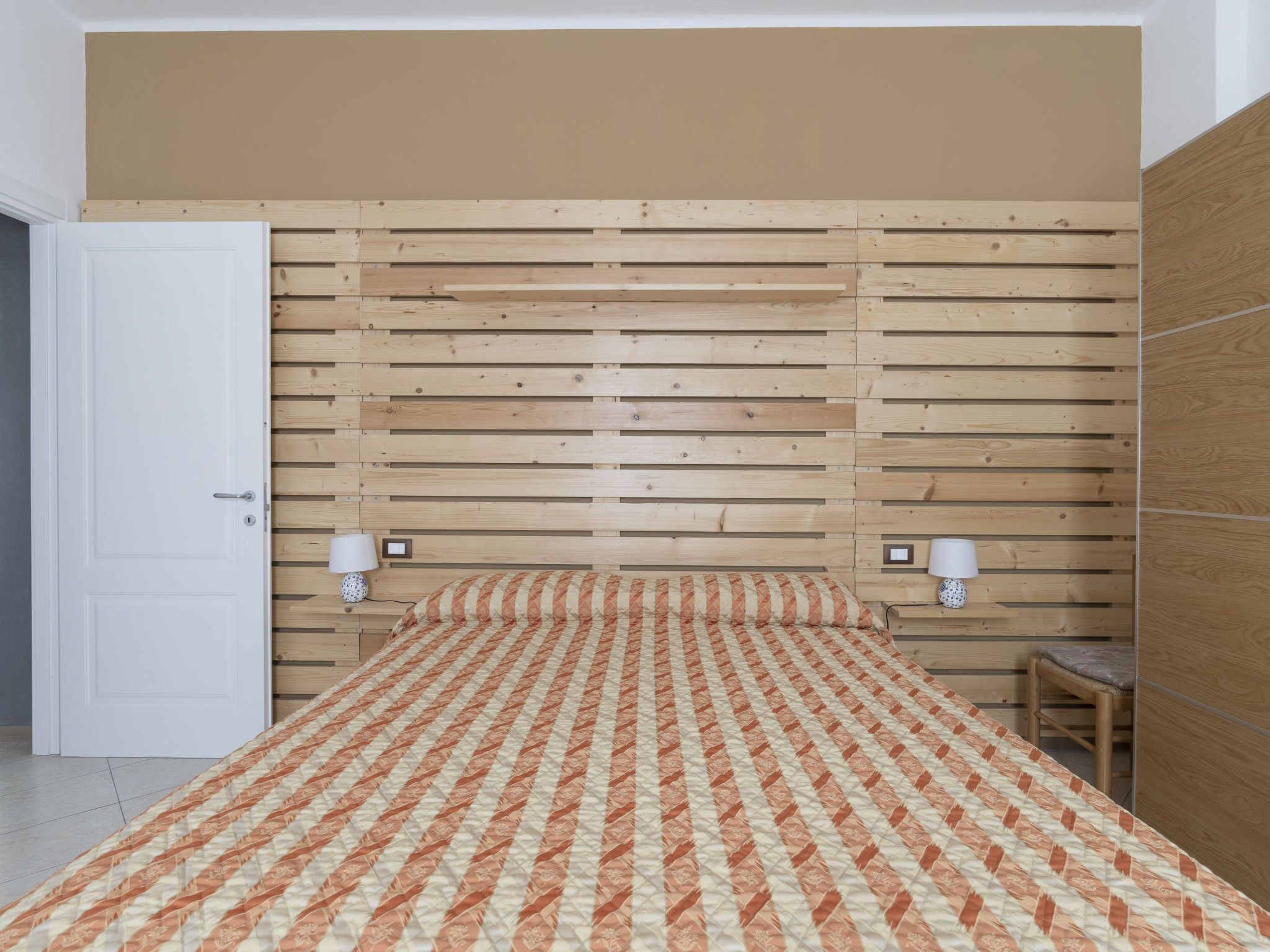 A bedroom with a wooden slat headboard, a bed with a patterned bedspread, two bedside lamps, and a wooden nightstand.