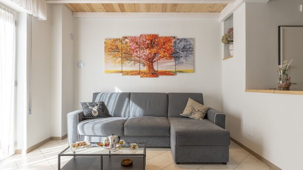 A modern living room with a grey sectional sofa, a glass coffee table with breakfast items, and a large autumn-themed painting on the wall.