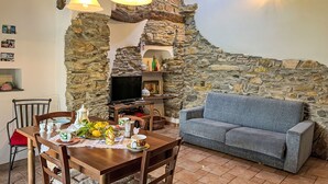 A cozy living room with a stone wall, a wooden dining table set with dishes, a grey sofa, and a television. - Ca' da Ciassa + Prima Porta (VLO132) by Interhome (Valloria)