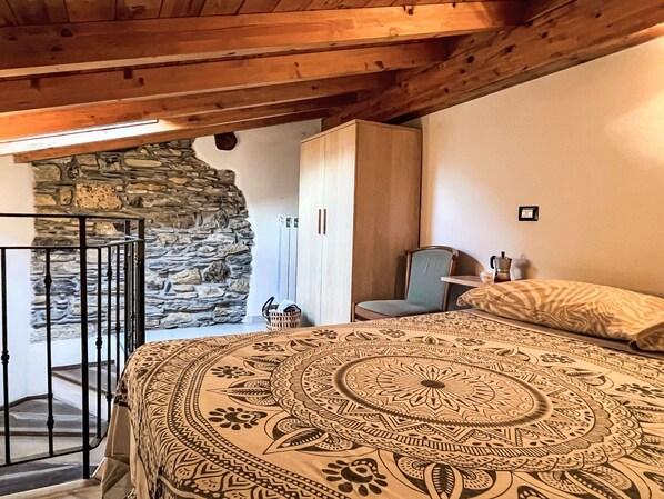 A bedroom with a stone wall, a bed with a patterned bedspread, a chair, a wardrobe, and a staircase. - Ca' da Prima Porta (VLO131) by Interhome (Valloria)
