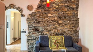 Une pièce chaleureuse avec un mur en pierre, un canapé bleu, une table recouverte d’une nappe à motifs floraux et une horloge au mur.