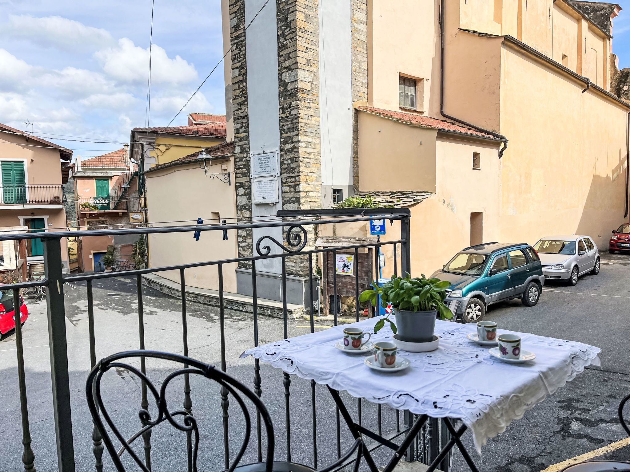 A balcony with a table set for tea, overlooking a street with parked cars and traditional buildings.
