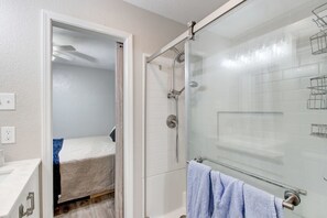 Un baño con cabina de ducha de vidrio, un lavabo blanco y una cama pequeña con una toalla azul.