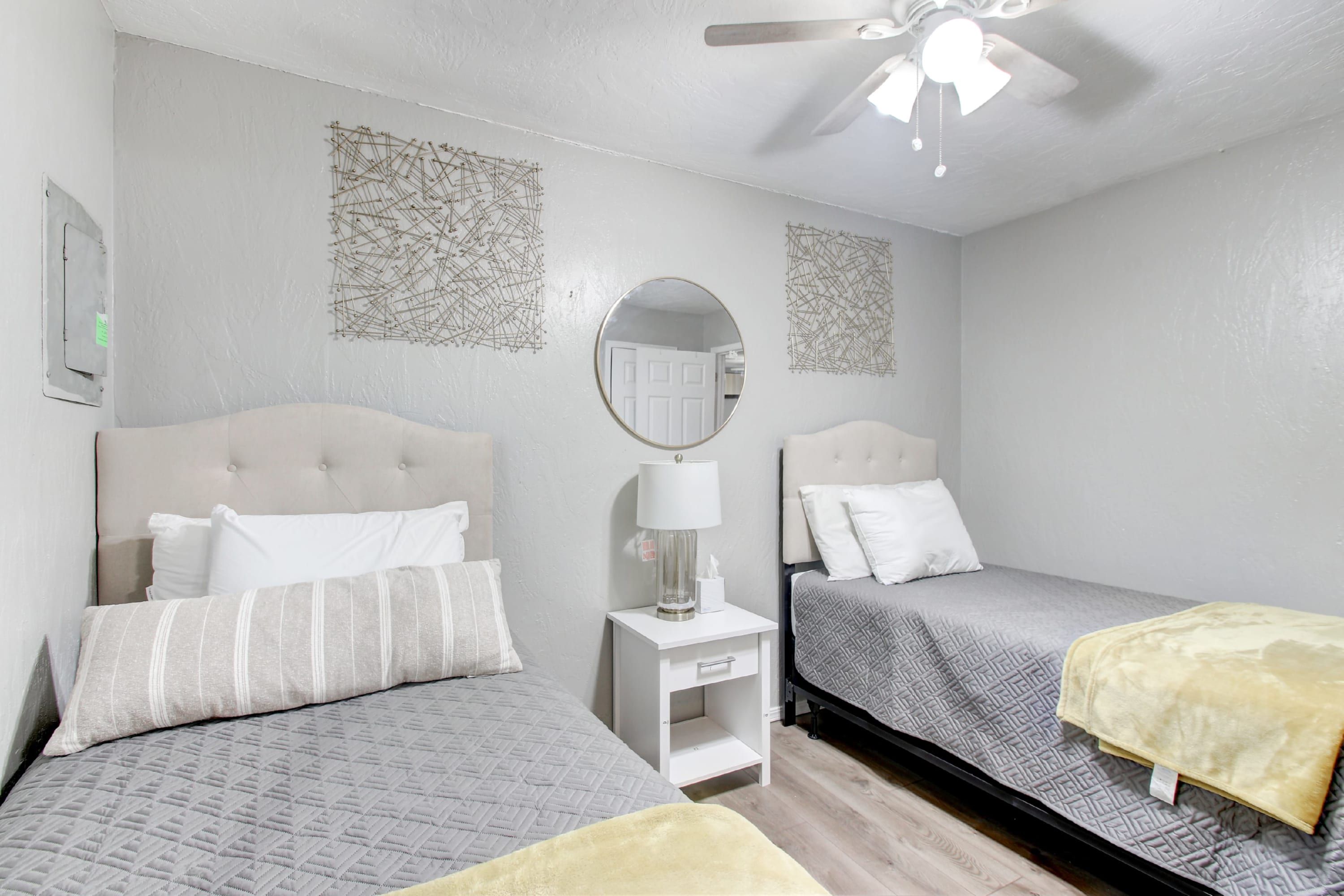 Une chambre d’hôtel avec deux lits, une petite table de chevet blanche, un miroir rond et un ventilateur de plafond.