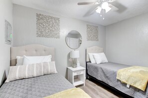 Habitación de hotel con dos camas, una mesita de noche pequeña y blanca, un espejo redondo y un ventilador de techo.