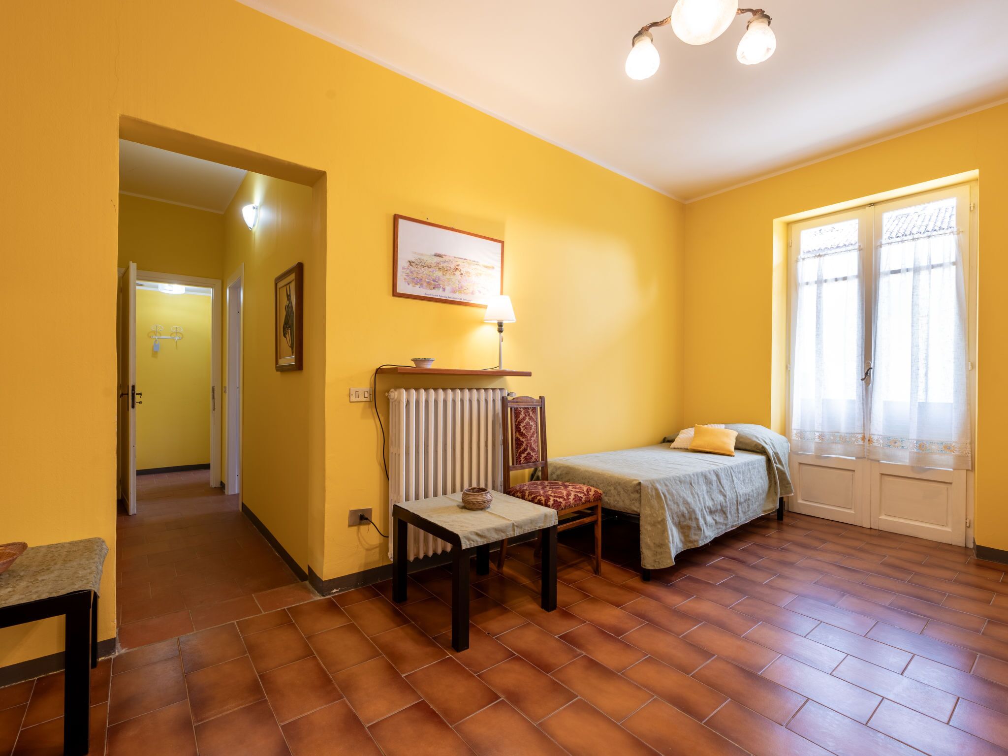 Ein Zimmer mit einem Bett, einem Tisch, einem Stuhl, einem Heizkörper und einem Vorhangfenster.