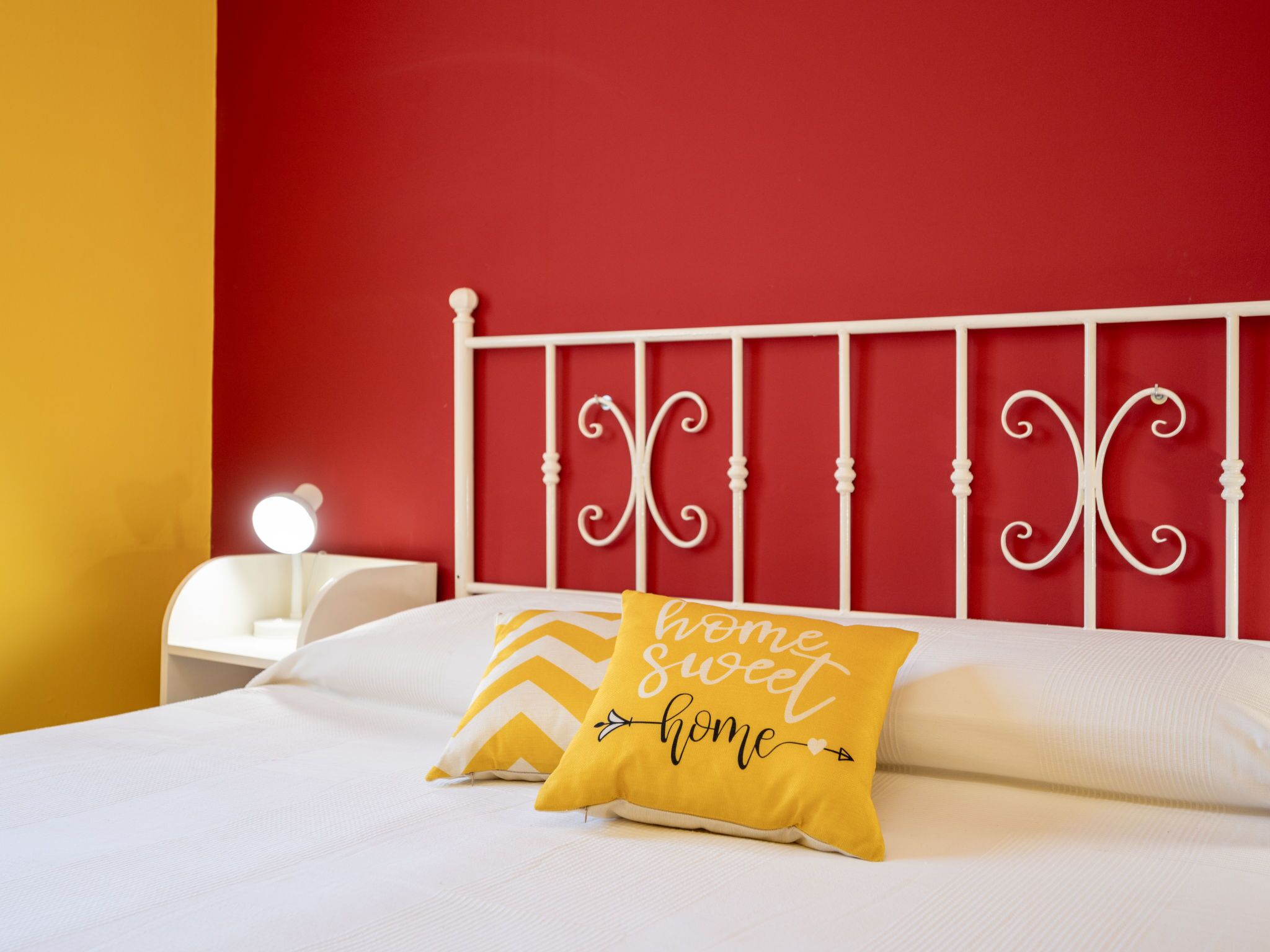 Una cama con ropa blanca, una almohada amarilla con el texto “hogar dulce hogar” y una pared roja.