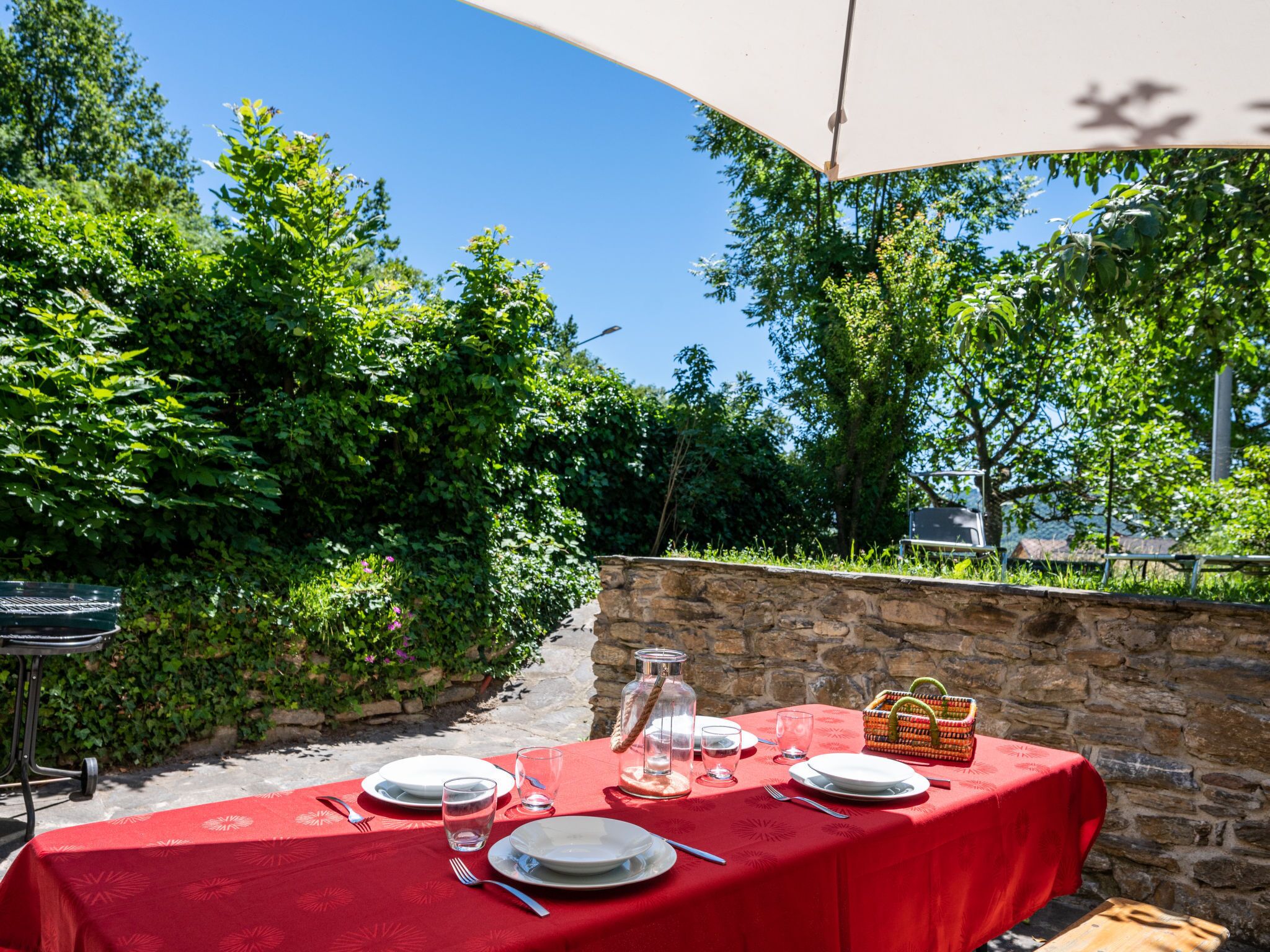 Un área de comedor al aire libre con un muro de piedra de fondo, un mantel rojo y una cesta de fruta.