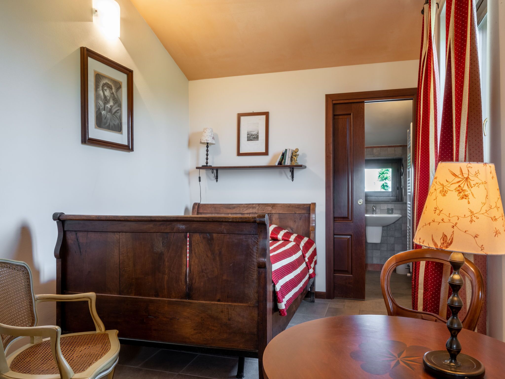 Un salon chaleureux avec un canapé en bois, une table à manger et une chaise. Un mur est orné d’une étagère où sont posés des livres et d’un tableau encadré. Une porte donne accès à une salle de bain.