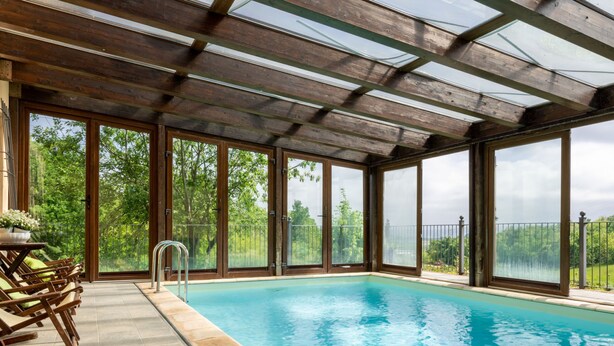 En swimmingpool med ett glastak, trädäck och utsikt över grönområden.