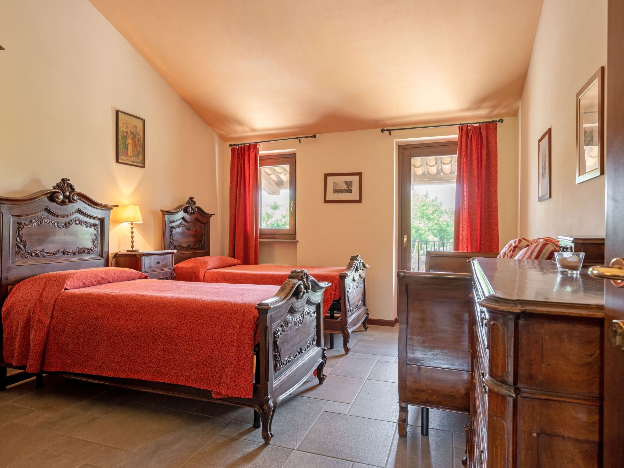 A hotel room with two beds, a wooden dresser, a desk, and a window with red curtains.
