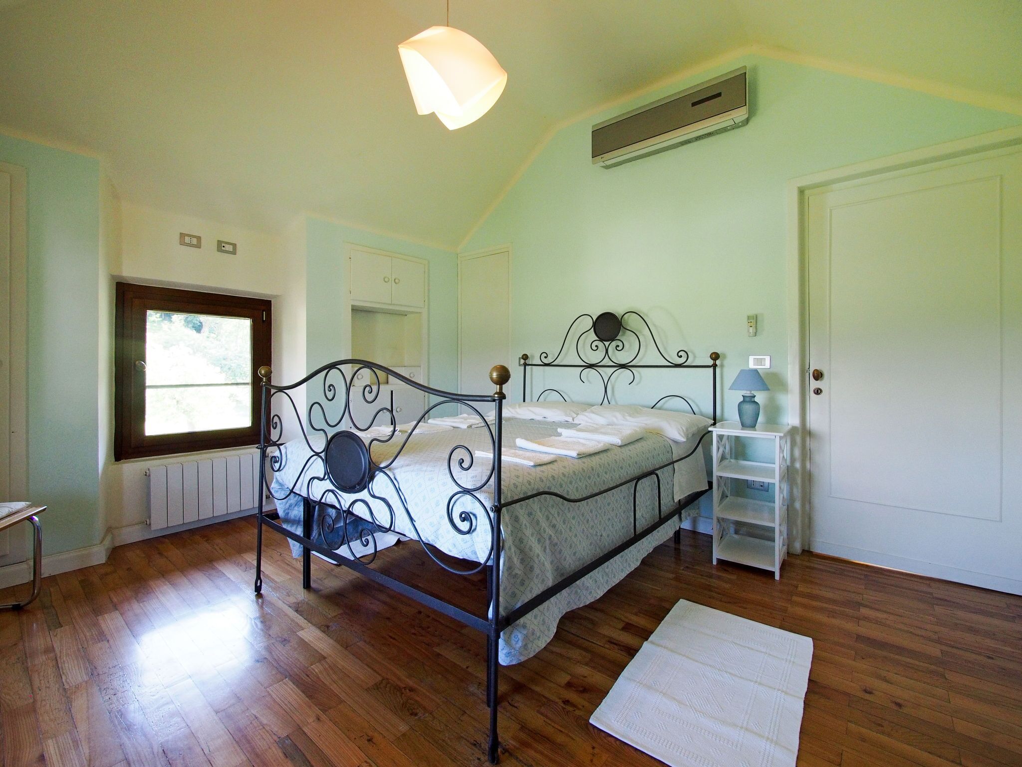 A bedroom with a metal bed frame, a window, a radiator, a white door, and a bedside table with a lamp.