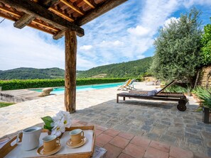 A poolside area with a wooden pergola, lounge chairs, and a table with a tray of cups and a pitcher.