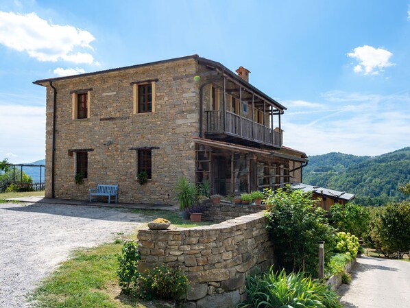 A stone building with a balcony, surrounded by greenery and a gravel driveway.