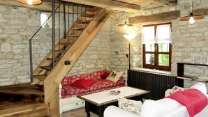 Ein rustikaler Raum mit einer Holztreppe, einer Steinsäule, einem roten, gemusterten Sofa, einem weißen Sessel und einem Tisch mit einer Schale.