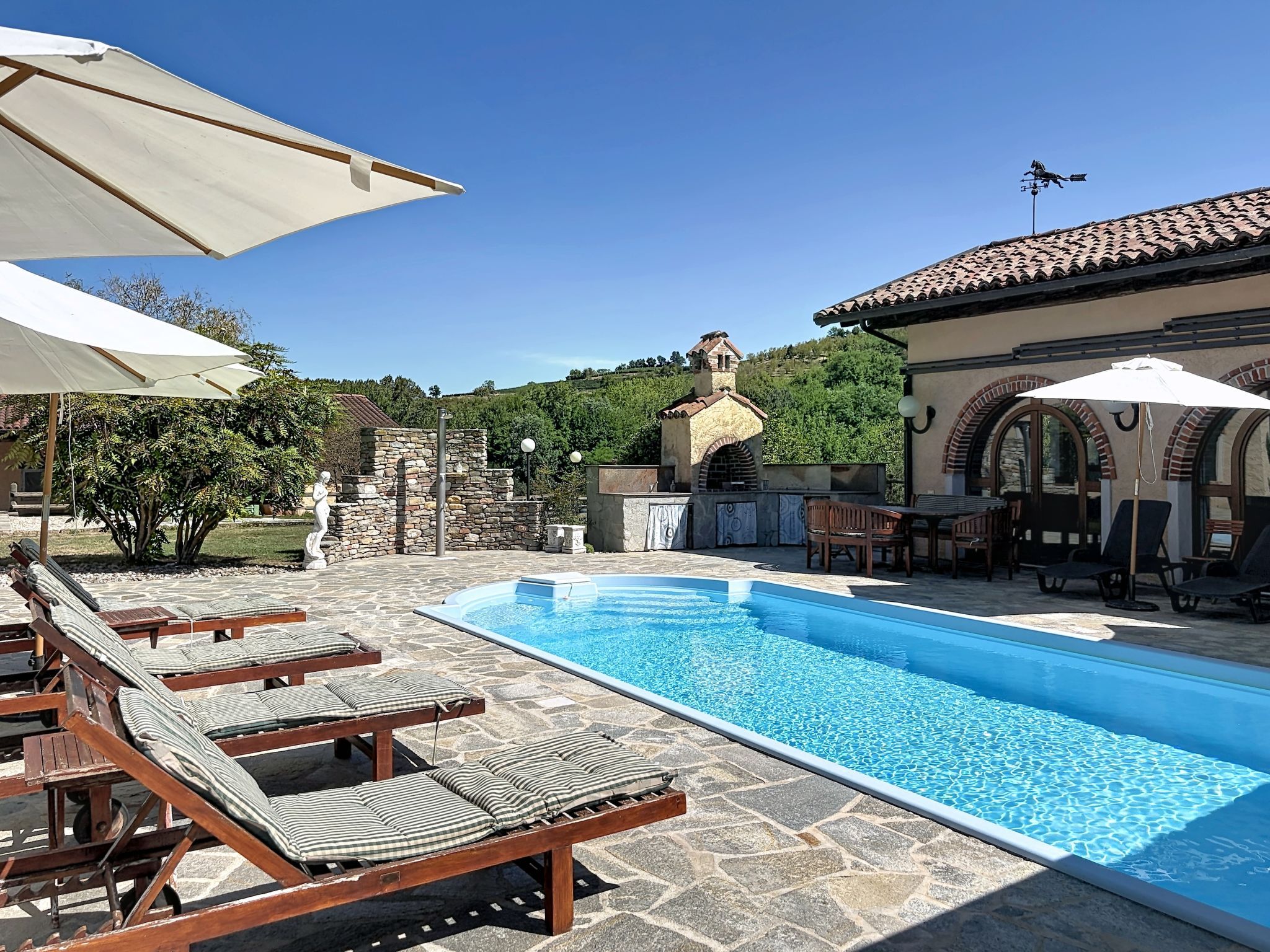 Piscina com espreguiçadeiras e guarda-sóis, parede de pedra e um edifício com telhado em azulejos.