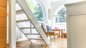 Un escalier en bois mène à une chambre située en mezzanine, offrant une vue sur les arbres grâce à de grandes fenêtres.