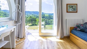 Une pièce avec un plancher en bois, un bureau blanc avec une chaise, un lit recouvert d’une couverture bleue et une vue sur un paysage verdoyant par une grande fenêtre.