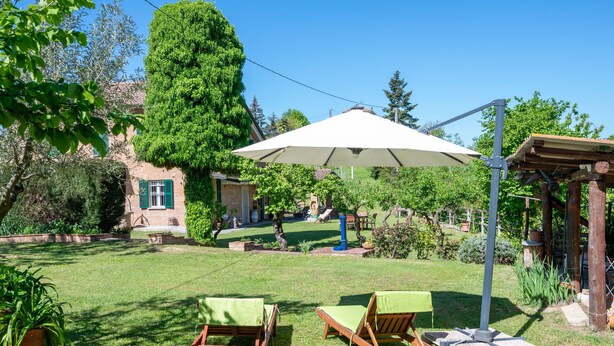 Ein Garten mit zwei Holzliegen, einer Pergola mit einem weißen Sonnenschirm und einem Backsteinhaus im Hintergrund.