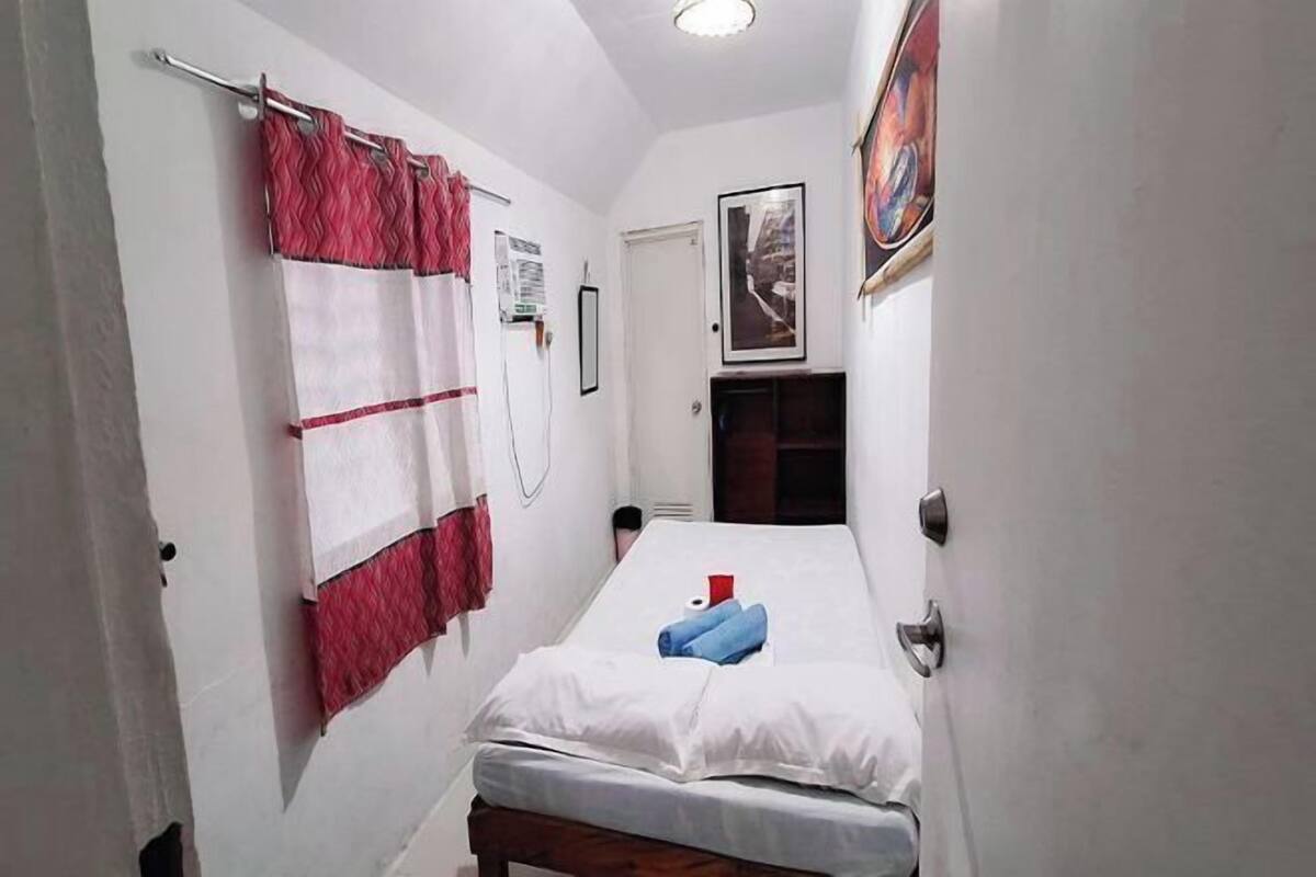 Ein schmales Zimmer mit einem Bett, einer Tür, einem Vorhang und einem gerahmten Bild an der Wand.