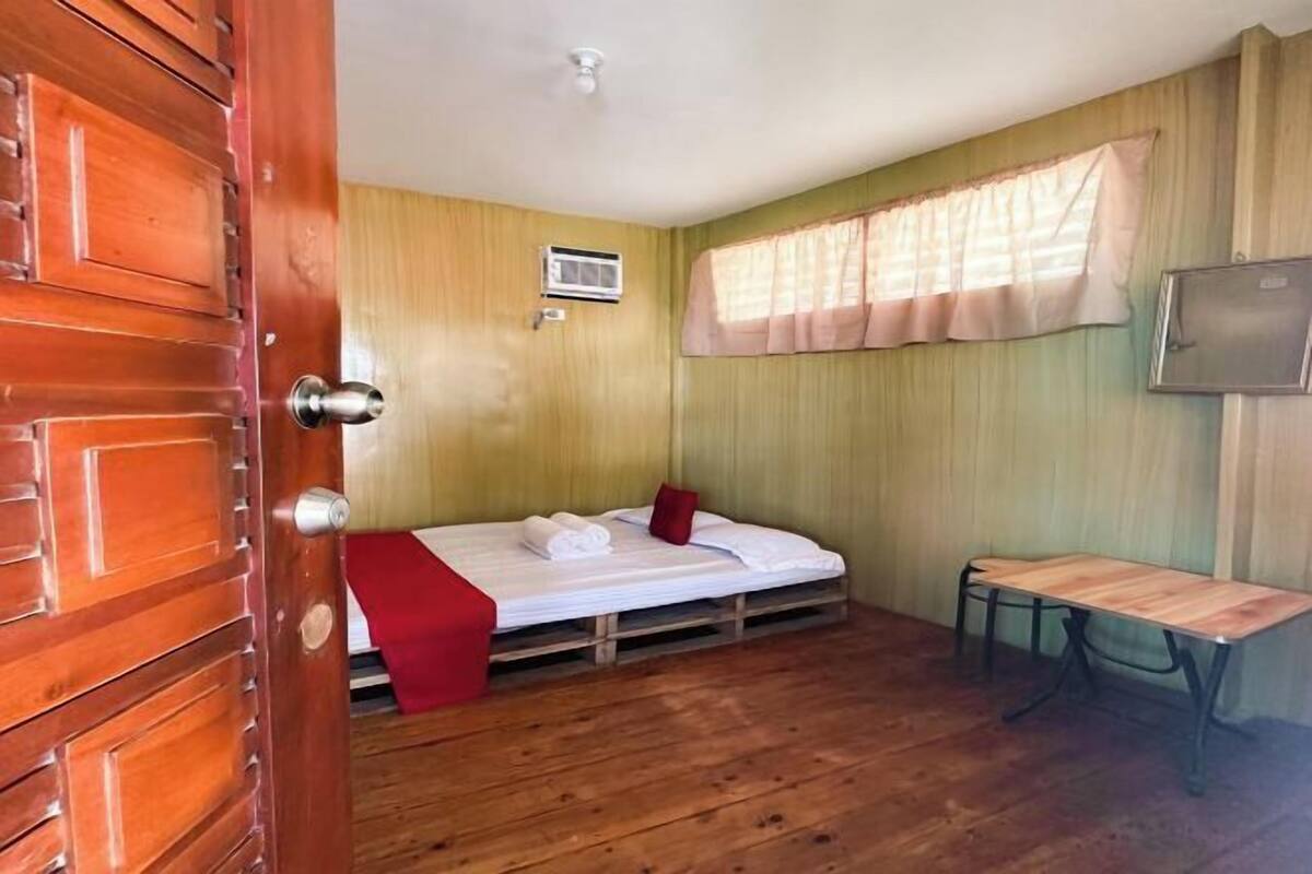 Ein Zimmer mit Holzboden, einem Bett mit weißer Bettw äsche, einem roten Kissen, einem kleinen Tisch, einem an der Wand montierten Fernseher und einem Fenster mit Vorhang.