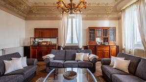 A living room with two sofas, a glass coffee table, and a chandelier. There is a kitchen area with wooden cabinets and a window with curtains. - Villa Loreto by Interhome (Costigliole d'Asti)