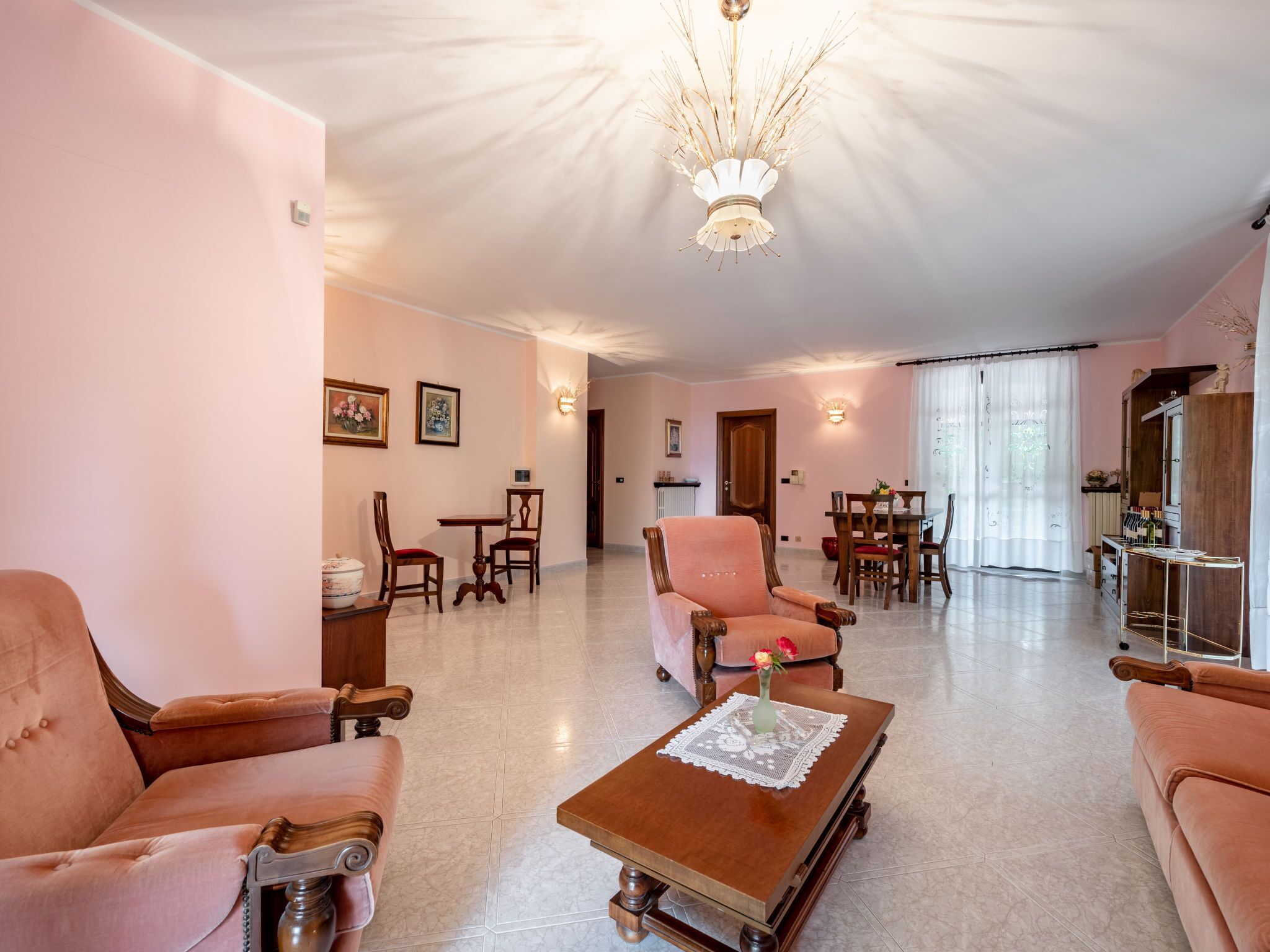 A spacious living room with a high ceiling, a chandelier, and a dining area with wooden furniture.