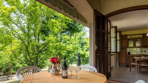 Zona d'exterior per dinar amb una taula preparada per a dues persones, envoltada d'idees blanques, sota un porxo cobert amb vista a arbres verds i frondosos.
