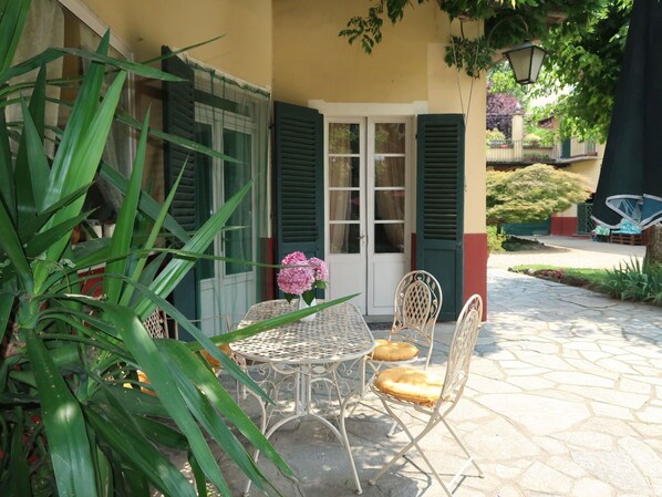 A patio with a round metal table and two chairs, a door with green shutters, and a potted plant in the foreground.