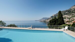 A clear blue swimming pool with lounge chairs, a view of the sea, and a hillside with buildings. - Parc Massolin by Interhome (Roquebrune Cap Martin)