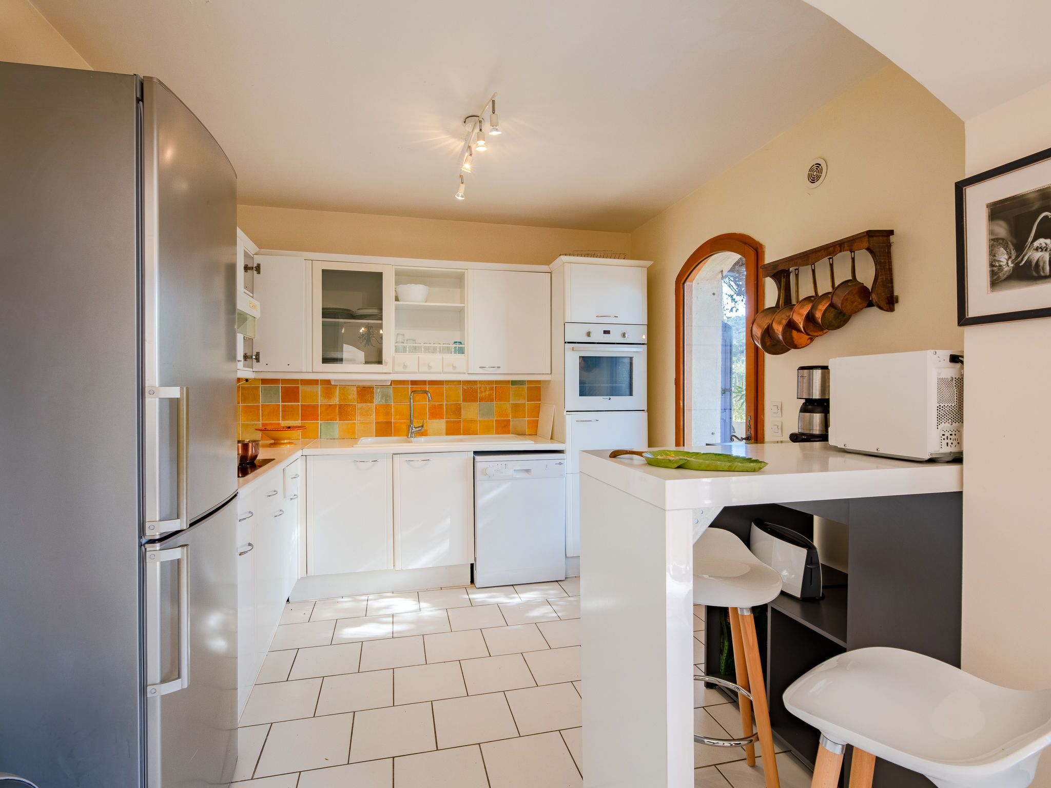 A modern kitchen with white cabinets, a stainless steel refrigerator, a dishwasher, and a small island with stools.