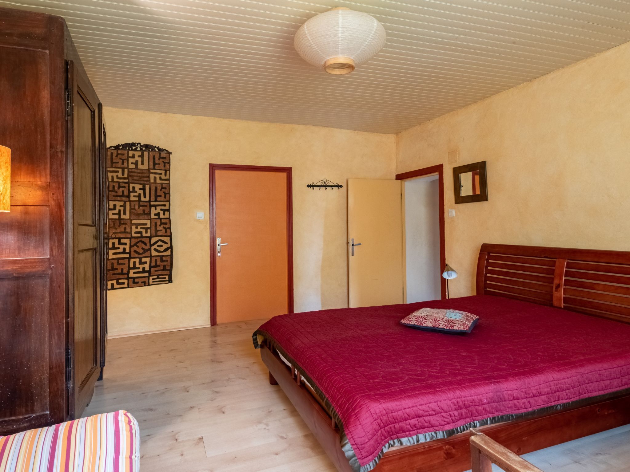 A bedroom with a wooden bed, a red bedspread, a wooden wardrobe, and a patterned tapestry on the wall.