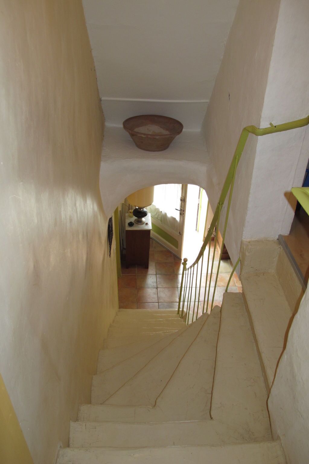 Un escalier étroit doté d’une rampe, menant à une pièce avec une fenêtre et une table sur laquelle se trouve une lampe.