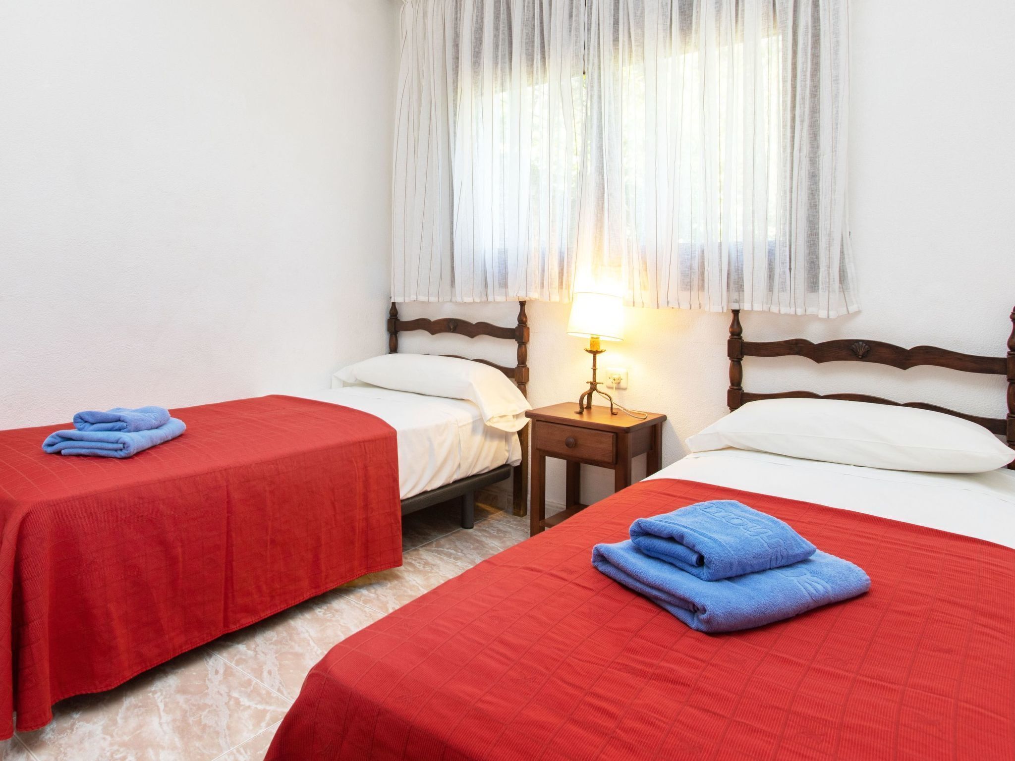 Quarto de hotel com duas camas de solteiro, cada uma com colcha vermelha e toalha azul. Há uma mesa de cabeceira com abajur entre as camas.