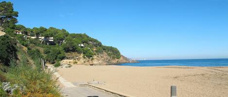 A beach with a paved walkway, a trash bin, and a bollard.