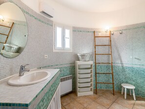 Une salle de bain avec un lavabo blanc, un miroir et des murs carrelés. On y trouve une fenêtre, un radiateur, une échelle et un petit tabouret.