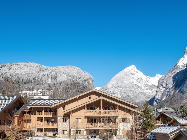 A multi-story wooden building with balconies, set against a backdrop of snow-covered mountains and clear blue sky.