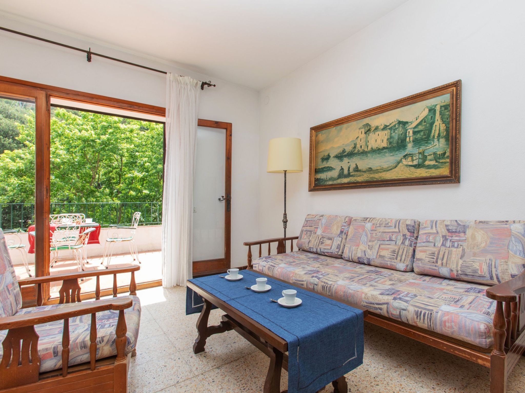A room with a wooden sofa, a dining table with a blue tablecloth, a painting on the wall, and a view of greenery outside.