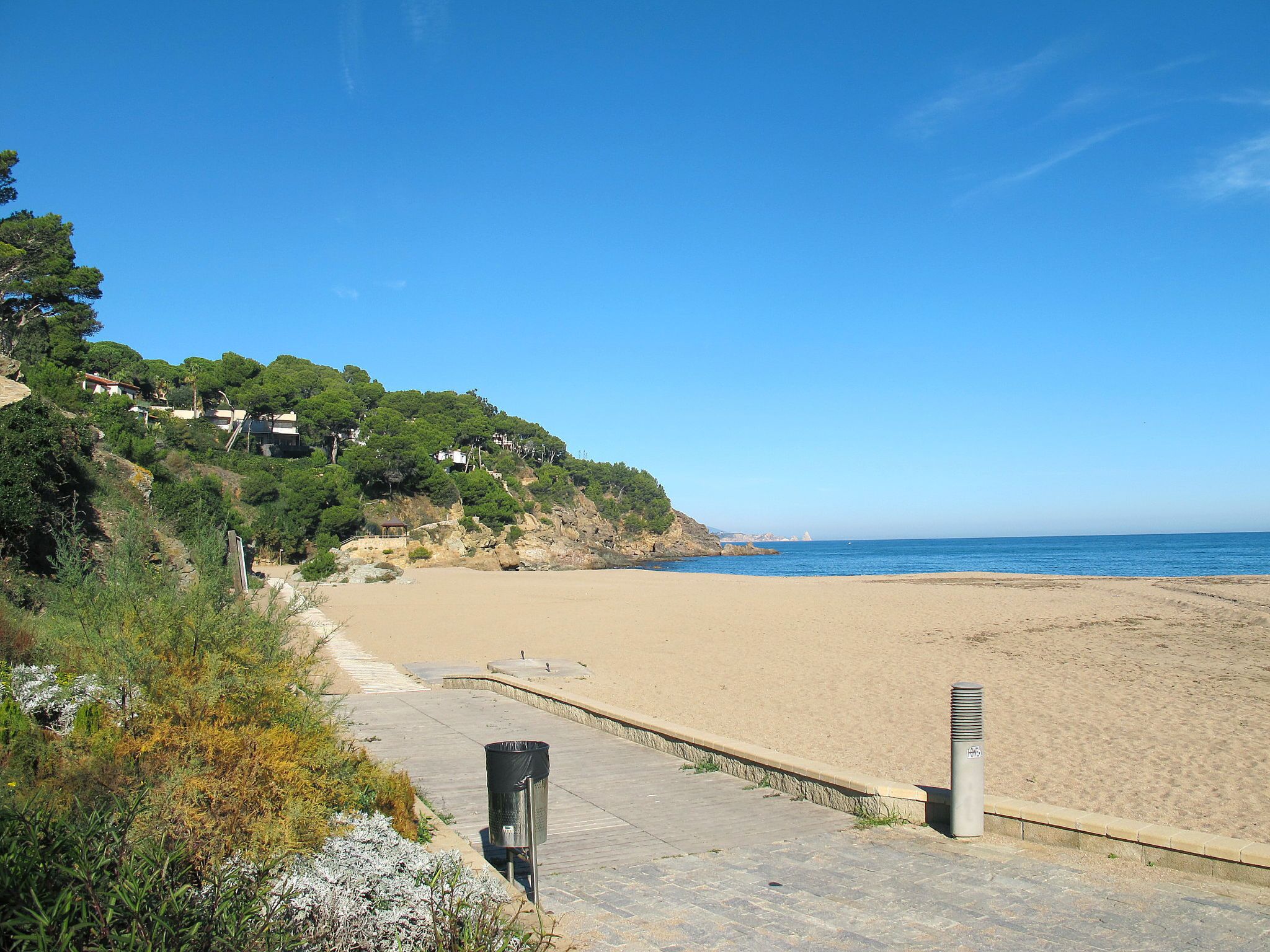 Une plage avec une promenade dallée, une poubelle et un poteau.
