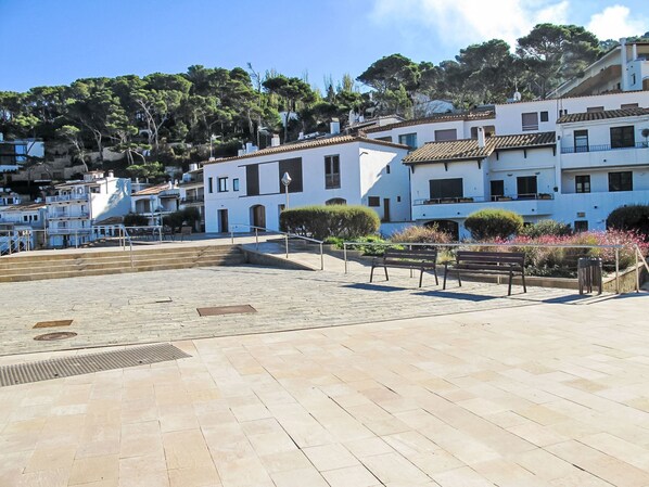 A paved square with benches and a small garden area, surrounded by white buildings with tiled roofs and balconies.