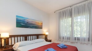 A hotel room with a bed, two bedside tables with lamps, a painting of a seascape, and a window with sheer curtains. - S'Esquirols Nº5 by Interhome (Sa Riera)