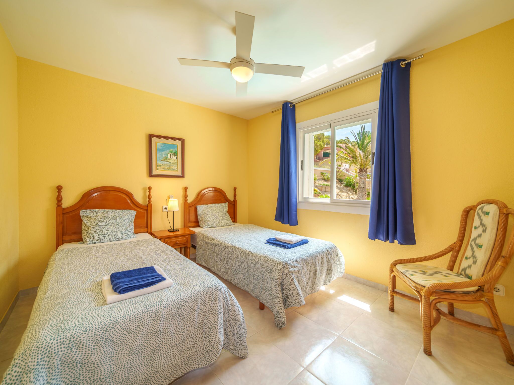 A hotel room with two beds, a ceiling fan, a window with curtains, a chair, and a painting on the wall.